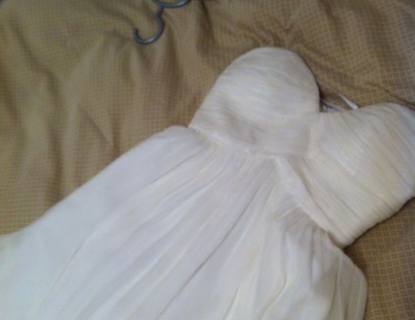 שמלת כלה לבנה עם סטרפלס פרושה על כיסוי מיטה בז', ומציגה את עיצוב הקפלים והבד הזורם. מתלה בגדים נראה חלקית ליד השמלה.