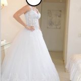 אדם בשמלת כלה לבנה ללא שרוולים עומדת בחדר מואר בעדינות. השמלה כוללת פרטי תחרה מורכבים על המחוך וחצאית מלאה ונשפכת. תפאורה בגוון ניטרלי נראה ברקע.