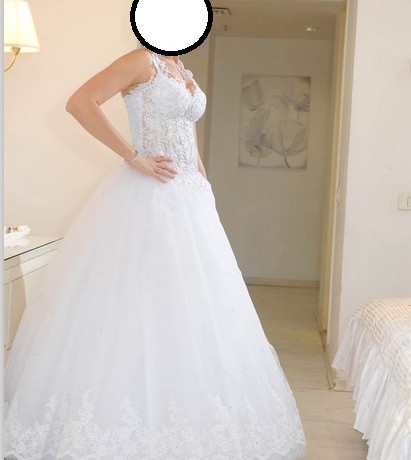 אדם בשמלת כלה לבנה ללא שרוולים עומדת בחדר מואר בעדינות. השמלה כוללת פרטי תחרה מורכבים על המחוך וחצאית מלאה ונשפכת. תפאורה בגוון ניטרלי נראה ברקע.