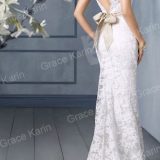 אישה בשמלת תחרה לבנה עם גב וי וקשת סאטן בז עומדת בחדר עם קירות אפורים ורצפת עץ. זר פרחים לבנים בחזית.