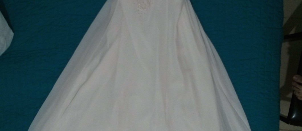 שמלת כלה לבנה עם חצאית מלאה ופרטי תחרה על המחוך פרושה על כיסוי מיטה צהבהב.