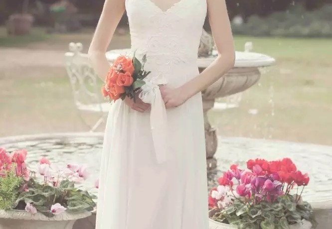 כלה בשמלה לבנה עומדת אוחזת בזר ורדים כתומים. היא נמצאת מול מזרקה, מוקפת פרחים אדומים וורודים. נראה שהתפאורה היא גן.