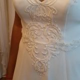 אדם שלובש שמלת כלה לבנה ללא שרוולים עם דפוסי פרחים רקומים מורכבים ועבודות חרוזים על המחוך, עומד בחדר כשברקע שמלות אחרות.