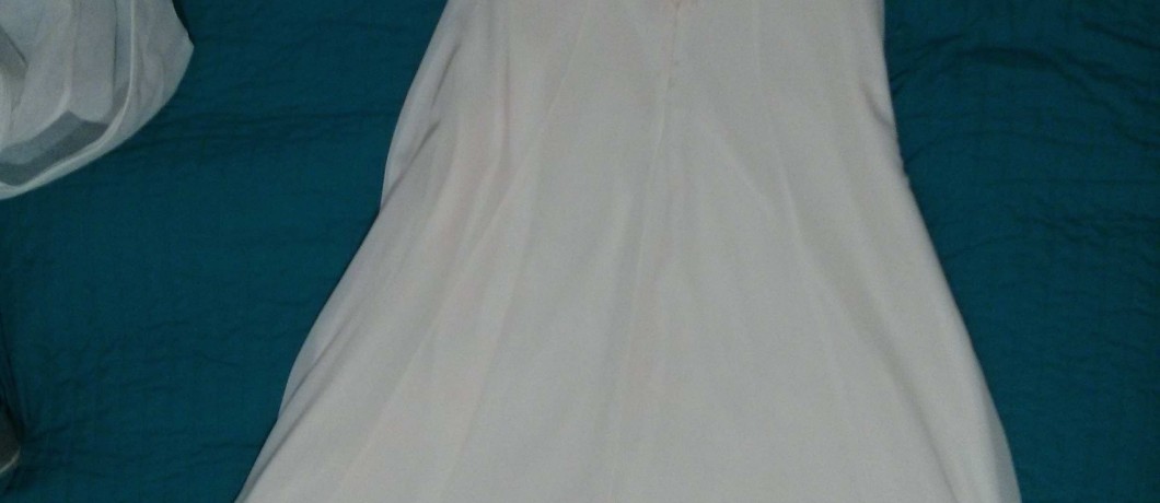 שמלה ורודה בהירה ללא שרוולים מונחת על שמיכה מרופדת בצבע ירוק כהה. לשמלה עיצוב זורם עם קו מותן מעט אסוף.