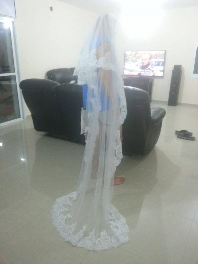 אדם לובש צעיף תחרה ארוך עומד בסלון עם רצפות אריחים. יש ספות עור שחורות וטלוויזיה ברקע. החדר נראה מואר.