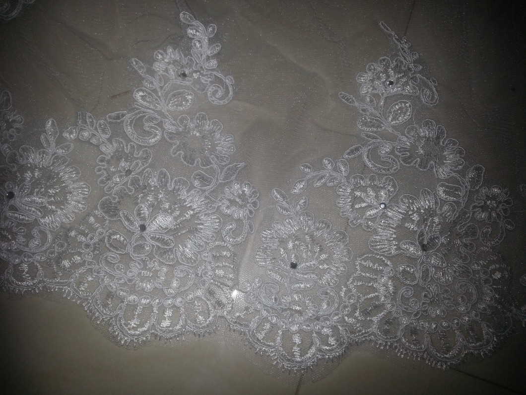 תקריב של בד תחרה לבן מורכב עם דוגמאות פרחוניות. העיצוב כולל פרחים בשכבות ומערבולות עדינות, המודגשות על ידי חרוזים קטנים ונוצצים. הבד מונח שטוח ומדגיש את האומנות העדינה.