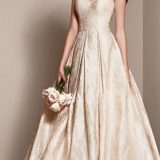 אישה בשמלת נשף אלגנטית ללא שרוולים שנהב עם דוגמה פרחונית. היא אוחזת בזר קטן של ורדים ורודים בהירים ועומדת על רקע נייטרלי, מציגה את החצאית המלאה של השמלה והמחוך המותאם.