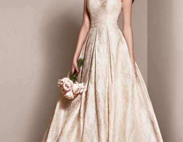 אישה בשמלת נשף אלגנטית ללא שרוולים שנהב עם דוגמה פרחונית. היא אוחזת בזר קטן של ורדים ורודים בהירים ועומדת על רקע נייטרלי, מציגה את החצאית המלאה של השמלה והמחוך המותאם.