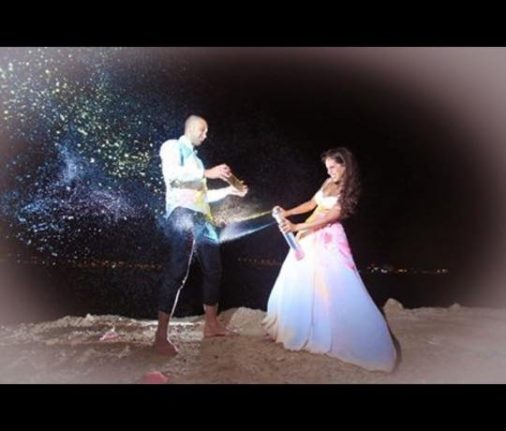 גבר ואישה עומדים על חוף הים בלילה, מתיזים אבקה צבעונית או נצנצים זה לכיוון השני. האישה לובשת שמלה לבנה, והגבר במכנסיים כהים וחולצה בהירה. הסצנה חגיגית ותוססת.