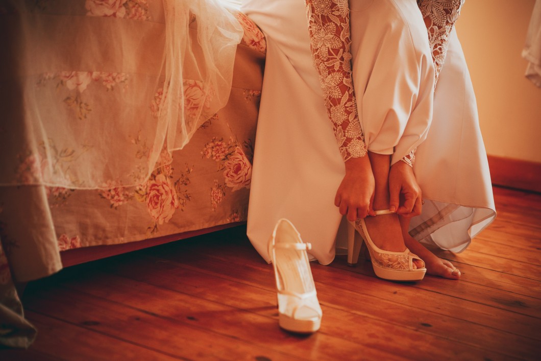 אדם בשמלת תחרה לבנה יושב על מיטה בדוגמת פרחים ומתאים נעל עקב. נעל אחת על, והשנייה על רצפת העץ הסמוכה. התפאורה חמה ואינטימית, מרמזת על רגע הכנה לאירוע.