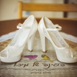 זוג נעלי עקב לבנות אלגנטיות עם הדגשות תחרה ורצועות קרסול ממוקמות על נייר דקורטיבי הכולל איורי ציפורים צבעוניים וטקסט בעברית. הרקע כולל כיסא עץ.