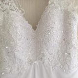 תקריב של שמלת כלה לבנה עם פירוט תחרה ופאייטים מורכבים, תלויה על קיר בצבע בהיר. התחרה יוצרת דוגמאות אלגנטיות על המחוך, ומוסיפה מגע של נצנוץ ואלגנטיות.