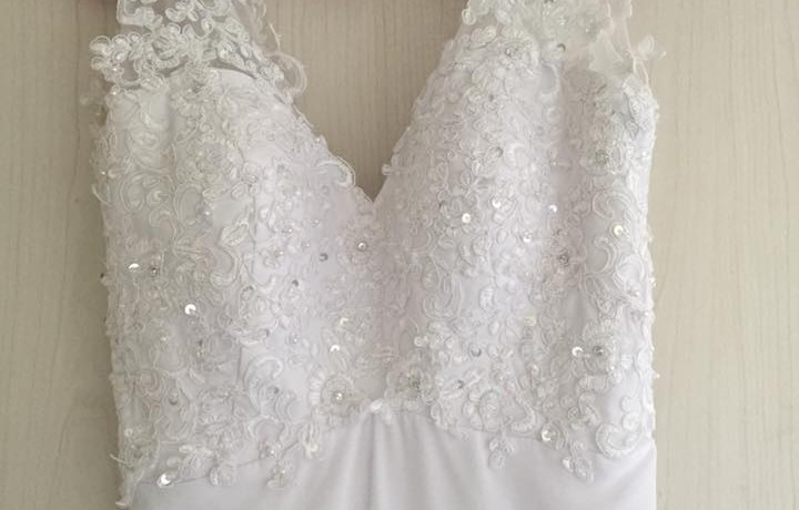 תקריב של שמלת כלה לבנה עם פירוט תחרה ופאייטים מורכבים, תלויה על קיר בצבע בהיר. התחרה יוצרת דוגמאות אלגנטיות על המחוך, ומוסיפה מגע של נצנוץ ואלגנטיות.