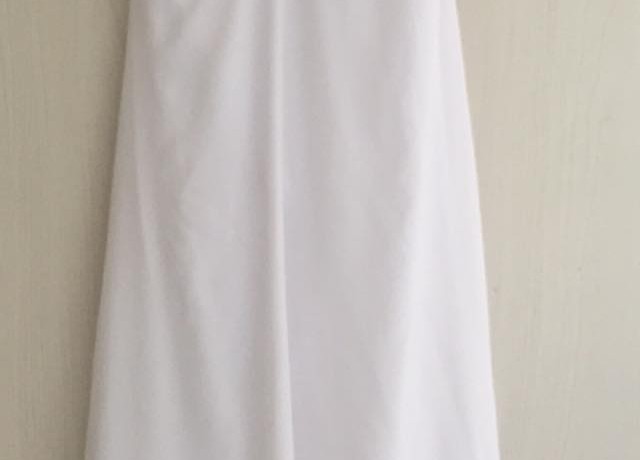 שמלה לבנה ללא שרוולים תלויה על קיר בהיר. השמלה כוללת מחוך מותאם וחצאית נשפכת וישרה.