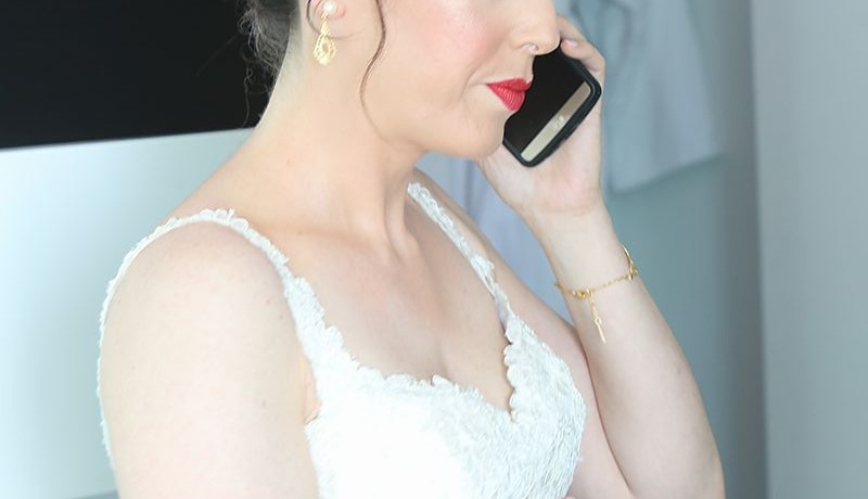אישה בשמלת תחרה לבנה מדברת בסמארטפון. יש לה שיער למעלה, לובשת שפתון אדום ועגילי זהב. זרועה השמאלית נראית, מראה צמיד. הרקע מטושטש בעדינות.