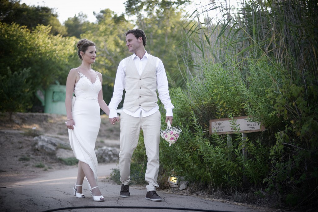 זוג לבוש בבגדי חתונה מחזיק ידיים תוך כדי הליכה בשביל מוקף בצמחייה. הכלה לובשת שמלה לבנה והחתן לובש גופיה קלה ומכנסיים. הוא מחזיק זר פרחים ביד אחת.