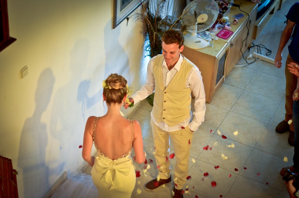 גבר ואישה עומדים בפנים על רצפת אריחים, מוקפים בעלי כותרת של ורדים. האישה, עם פרח בשיער, פונה הצידה, לובשת שמלה. האיש, מחייך, מושיט ורד. אחרים נמצאים בקרבת מקום, נראים חלקית.