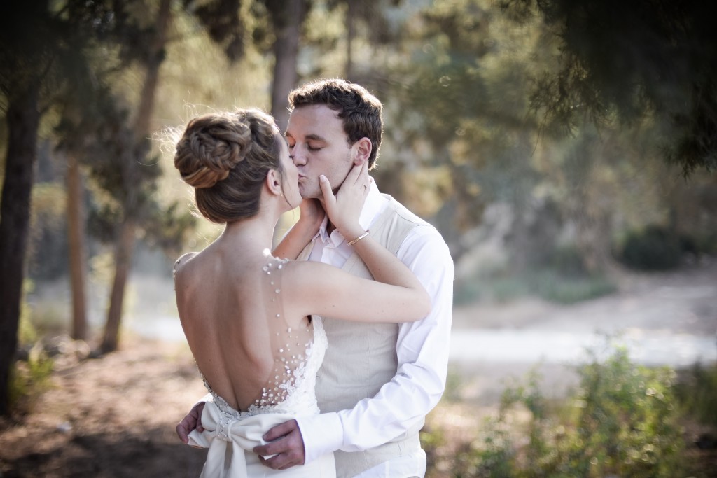 זוג חולק נשיקה בחוץ, מוקף בעצים. האישה לובשת שמלת כלה ללא גב עם פרטים פרחוניים עדינים, והגבר לבוש בחולצה לבנה עם אפוד בז. הסצנה מוארת ברכות, יוצר אווירה רומנטית.