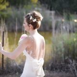 כלה בשמלה לבנה עם פרטים פרחוניים בשערה עומדת בחוץ ליד בריכה. היא מחזיקה זר פרחים ורוד ולבן ויש לה שמלת גב פתוחה עם קשת. דשא גבוה וירק מקיפים את הסצנה.