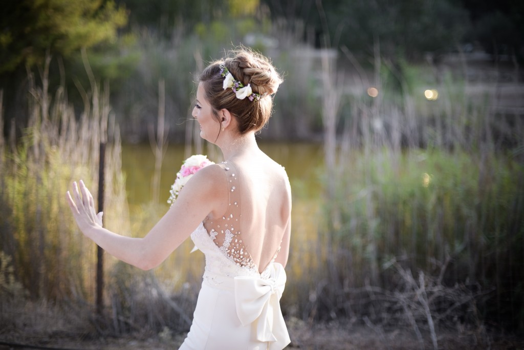 כלה בשמלה לבנה עם פרטים פרחוניים בשערה עומדת בחוץ ליד בריכה. היא מחזיקה זר פרחים ורוד ולבן ויש לה שמלת גב פתוחה עם קשת. דשא גבוה וירק מקיפים את הסצנה.