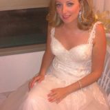 אישה בשמלת כלה תחרה לבנה יושבת בתוך הבית. לשמלה רצועות תחרה עדינות וחצאית מלאה. יש לה שיער גלי עד הכתפיים והיא מחייכת בעדינות, כשידיה מונחות על ברכיה.