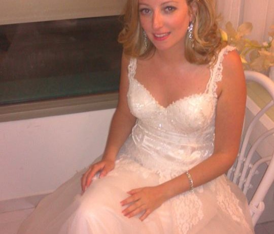 אישה בשמלת כלה תחרה לבנה יושבת בתוך הבית. לשמלה רצועות תחרה עדינות וחצאית מלאה. יש לה שיער גלי עד הכתפיים והיא מחייכת בעדינות, כשידיה מונחות על ברכיה.