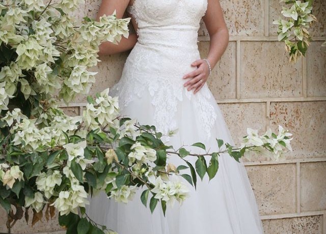 כלה בשמלת כלה תחרה לבנה עומדת על קיר אבן. פרחים לבנים דקורטיביים נמצאים בחזית. ידה הימנית מונחת על מותנה, וידה השמאלית מונחת בעדינות על מותניה.