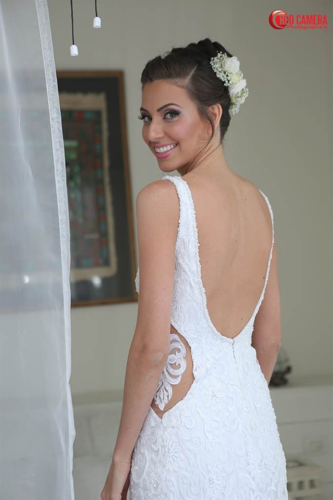 אישה בשמלת תחרה לבנה עם גב פתוח, מעוטרת בפרחים בשערה, מחייכת כשהיא עומדת ליד וילון שקוף. תמונה ממוסגרת תלויה על הקיר ברקע.