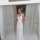 אישה חייכנית בשמלת תחרה לבנה עומדת מחזיקה וילונות שקופים פתוחים. יש לה פרח בשיער והיא עומדת בחדר מואר עם יצירות אמנות מעל.