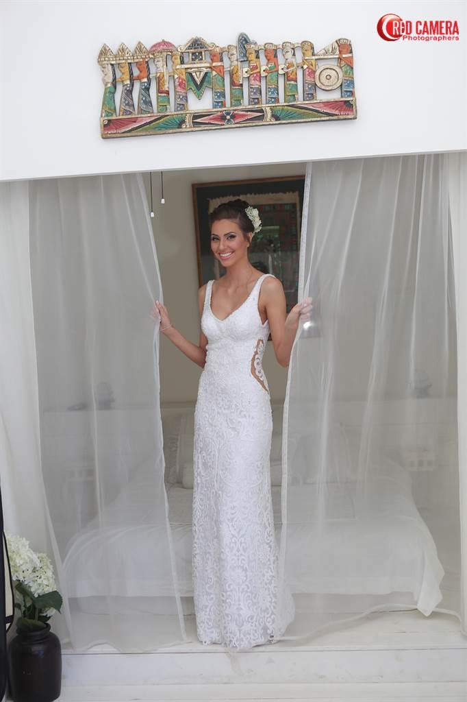 אישה חייכנית בשמלת תחרה לבנה עומדת מחזיקה וילונות שקופים פתוחים. יש לה פרח בשיער והיא עומדת בחדר מואר עם יצירות אמנות מעל.