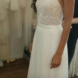 אדם לובש שמלת כלה לבנה תחרה ושיפון ללא שרוולים, עומד בתוך הבית. לשמלה טופ תחרה פרחוני עם רצועות ספגטי וחצאית שיפון נשפכת. חלק מאדם אחר נראה ברקע.