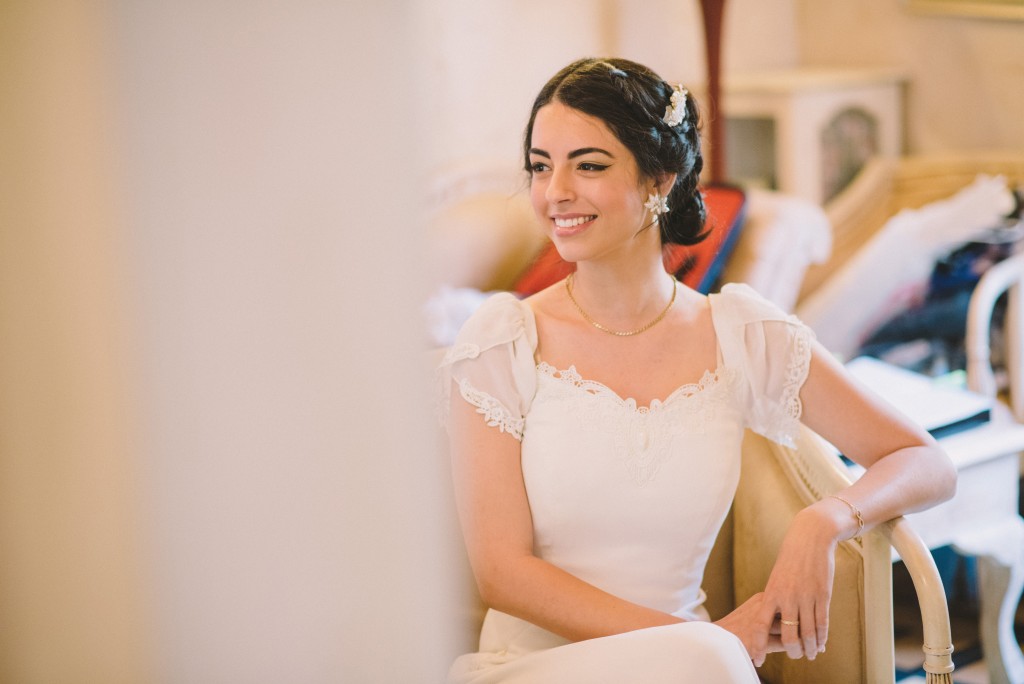 אישה בשמלת כלה לבנה יושבת על כיסא ומחייכת. יש לה שיער כהה מעוצב באלגנטיות, מעוטר בתספורת קטנה. הרקע מציג גוונים רכים ונייטרליים ורהיטים, היוצרים אווירה חמימה.