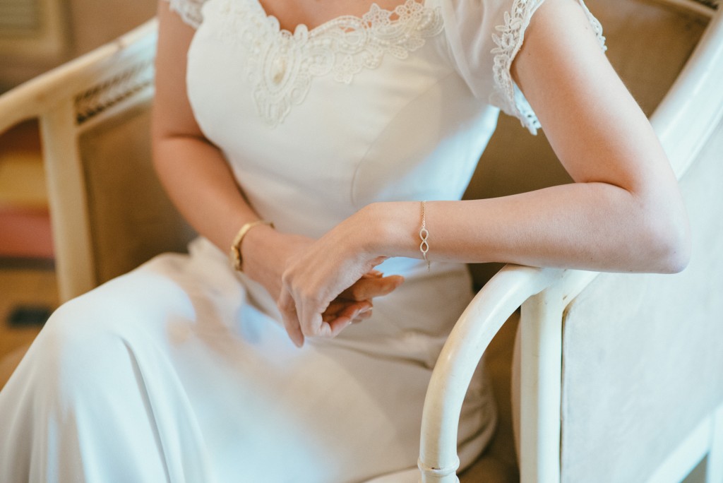 אישה בשמלה לבנה עם פרטי תחרה יושבת על כיסא בצבע שמנת. היא עונדת צמיד עדין וצמיד. זרועותיה מונחות על משענות הכיסא.