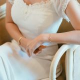 אדם יושב בשמלה לבנה מכוסת תחרה מניח את זרועותיו על משענת כיסא. צמיד ושעון זהב מעטרים את פרקי ידיהם. התמונה לוכדת רגע אלגנטי ושליו המתמקד בפרטי השמלה והתכשיטים.