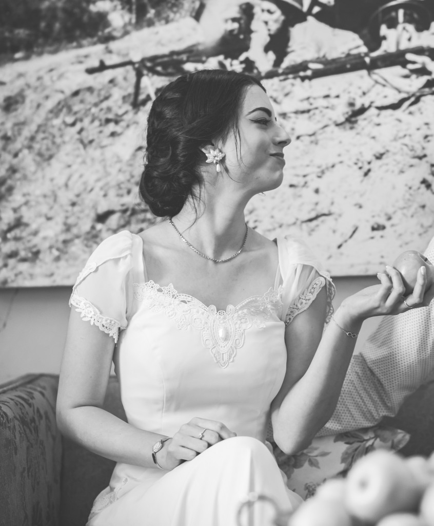 אישה בשמלה בסגנון וינטג יושבת על ספה, מחייכת בעיניים עצומות, מחזיקה תפוח. שיערה מעוצב באלגנטיות, והיא עונדת עגיל פרחים. ברקע יש תמונה גדולה בשחור-לבן. התמונה בשחור לבן.