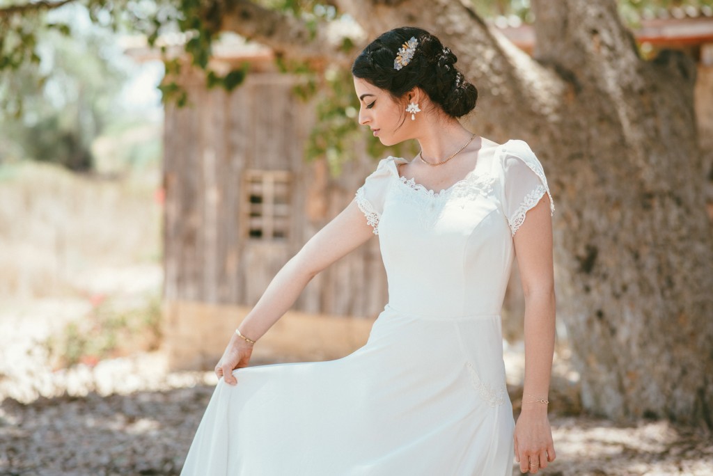 אישה בשמלה לבנה עומדת בחוץ, אוחזת בשולי שמלתה הגולשת. היא עונדת פריט שיער פרחוני ותכשיטים. ברקע מבנה עץ כפרי ועץ גדול. התפאורה נראית שטופת שמש וטבעית.