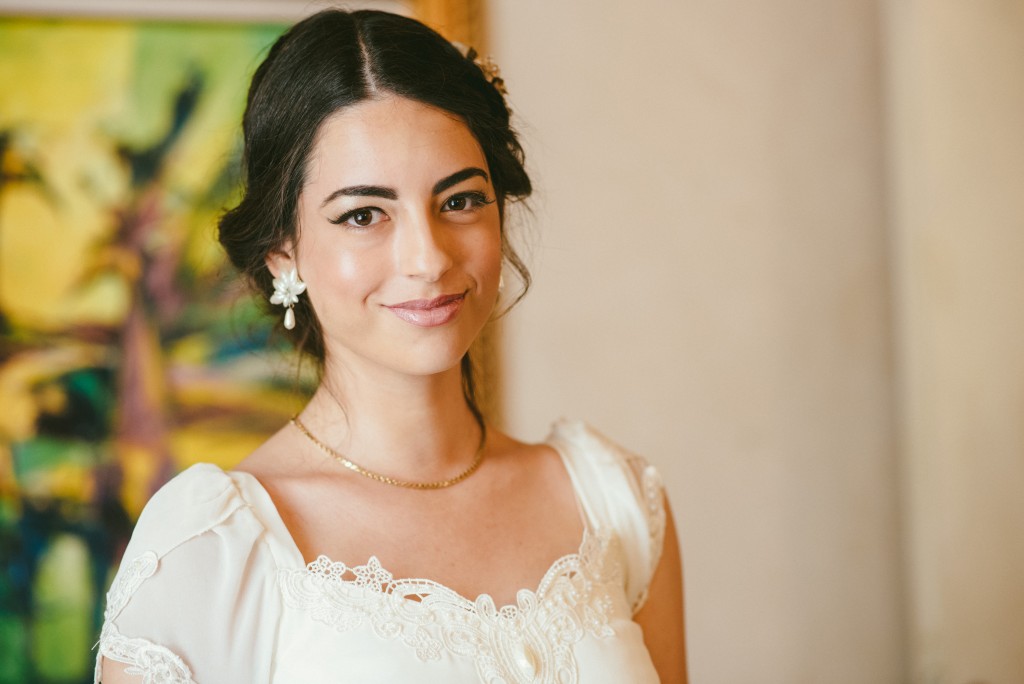 אישה עם שיער כהה ארוך מעוצב באפוד לובשת שמלה לבנה עם פרטי תחרה. היא מחייכת קלות ופונה למצלמה. ציור צבעוני מטושטש ברקע.