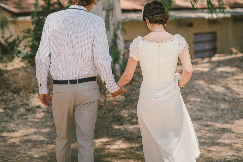 זוג מחזיק ידיים בזמן הליכה בחוץ ביום שמש. האישה לובשת שמלה לבנה והגבר בחולצה בהירה ומכנסיים. ברקע עצים גבוהים ומבנה כפרי.