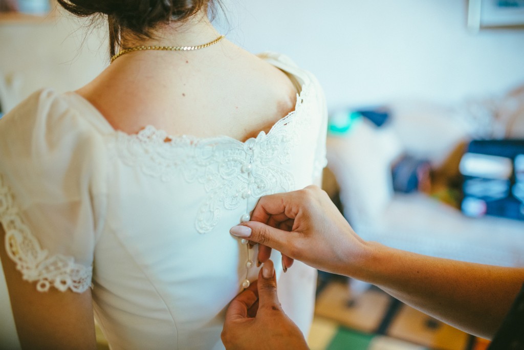 אדם מהדק כפתורים בגב שמלת כלה לבנה מעוטרת תחרה שלבשה על ידי אדם אחר. הסצנה היא בתוך הבית, עם תאורה רכה, המתמקדת בפרטי התחרה המורכבים ובידיים מהדקות את השמלה.