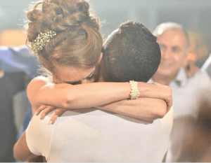 אישה עם שיער מעוצב וחתיכת שיער מחבקת גבר בחולצה לבנה. זרועותיה כרוכות סביבו בחוזקה. אדם נוסף ברקע מחייך, מעט מטושטש. הסצנה משדרת רגש וקרבה.