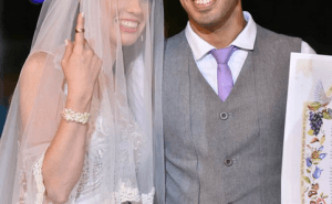 חתן וכלה מחייכים ועומדים יחד. הכלה לובשת שמלה והינומה לבנה, והחתן בגופייה אפורה עם עניבה סגולה. הם אוחזים במסמך מעוטר.