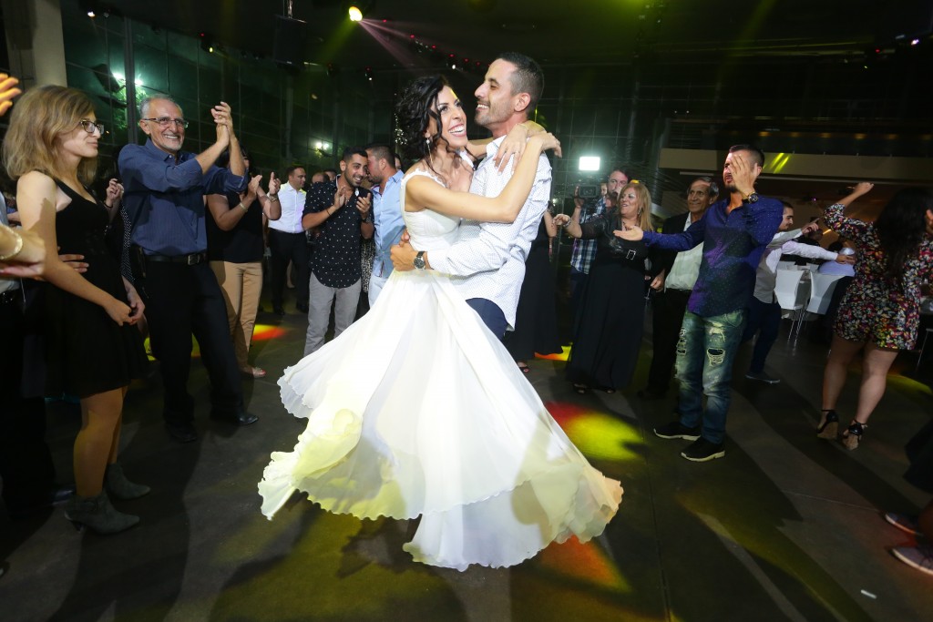 חתן וכלה רוקדים בשמחה בקבלת הפנים לחתונה שלהם, מוקפים באורחים מחייכים. שמלתה הלבנה של הכלה מסתחררת סביבם, ואורות צבעוניים יוצרים אווירה תוססת על רחבת הריקודים.