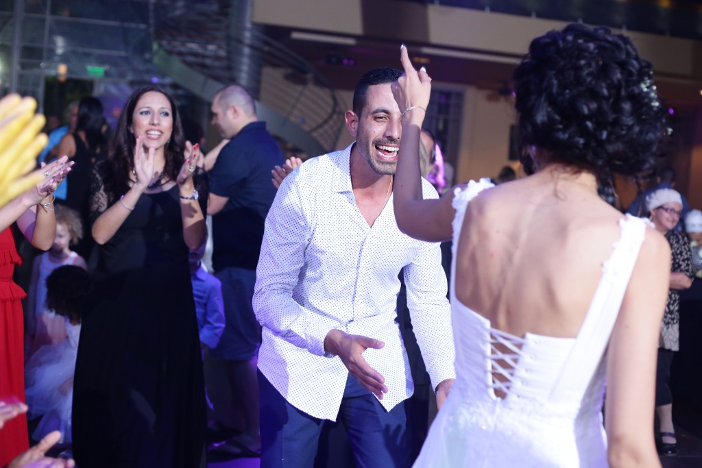 גבר בחולצה לבנה מחייך ורוקד עם אישה בשמלה לבנה באירוע תוסס. הם מוקפים באנשים אחרים מוחאים כפיים ומריעים באווירה חגיגית עם תאורה עמומה.