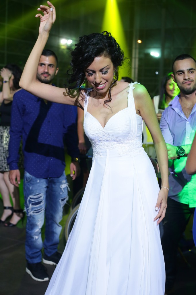 אישה בשמלה לבנה רוקדת בשמחה, מוקפת באנשים במסיבה תוססת עם אורות בהירים. היא מחייכת ומרימה זרוע אחת, כשאחרים לבושים בלבוש לא פשוט נהנים מהרגע.