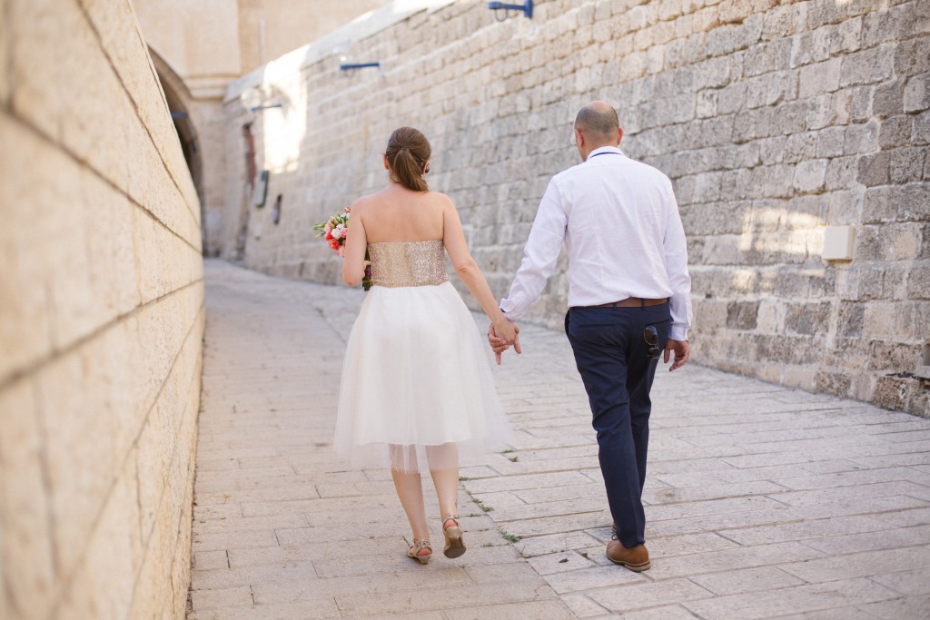 זוג הולך יד ביד בסמטה צרה ומרוצפת אבן. האישה לובשת שמלה לבנה ומחזיקה זר, בעוד הגבר בחולצה לבנה ומכנסיים כהים. אור השמש מטיל צללים רכים על הקירות, מה שמרמז על יום חמים ושטוף שמש.