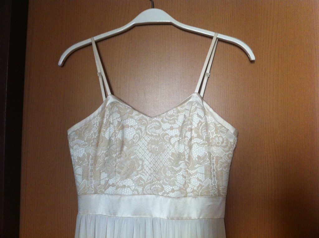 שמלה לבנה עם כתפיות דקות ומחוך בדוגמת תחרה תלויה על קולב לבן על רקע עץ. התחרה מציגה עיצובים פרחוניים, ולשמלה רצועת סאטן במותניים.