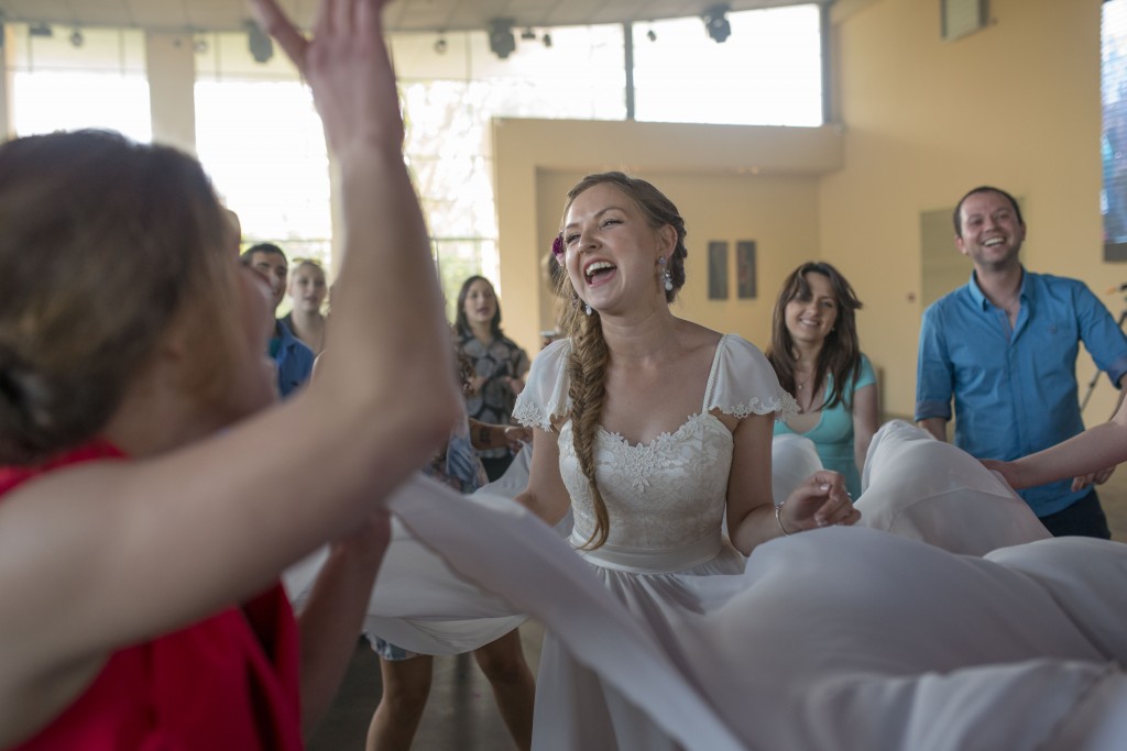 כלה חייכנית עם צמה ופרח בשיער רוקדת בשמחה עם אורחים במקום מואר ומקורה. אנשים סביבה, כולל אישה באדום וגבר בכחול, גם הם מחייכים וחוגגים.