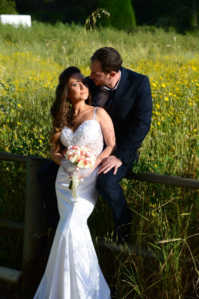 כלה בשמלה לבנה אוחזת בזר ויושבת על גדר בשדה של פרחים צהובים. גבר בחליפה כהה יושב לידה, מביט בה. הרקע שופע בירק.