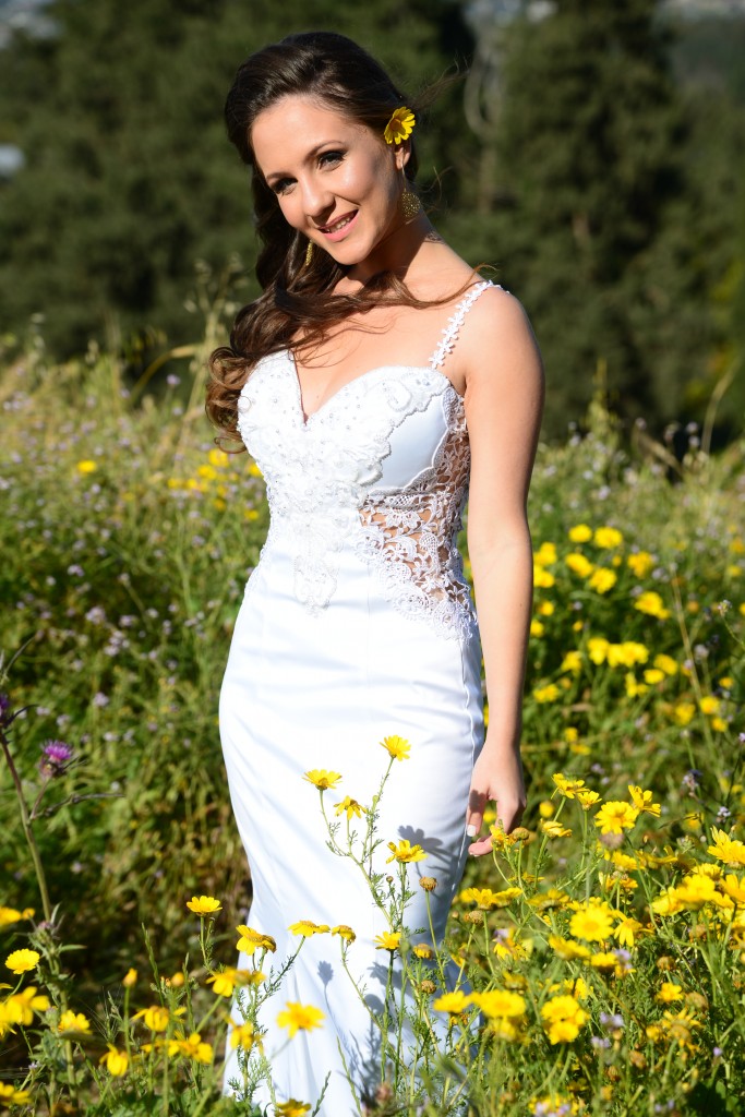 אישה בשמלת תחרה לבנה עומדת בשדה של פרחים צהובים. יש לה שיער חום ארוך עם פרח צהוב תחוב מאחורי אוזנה והיא מחייכת על רקע עצים ירוקים.