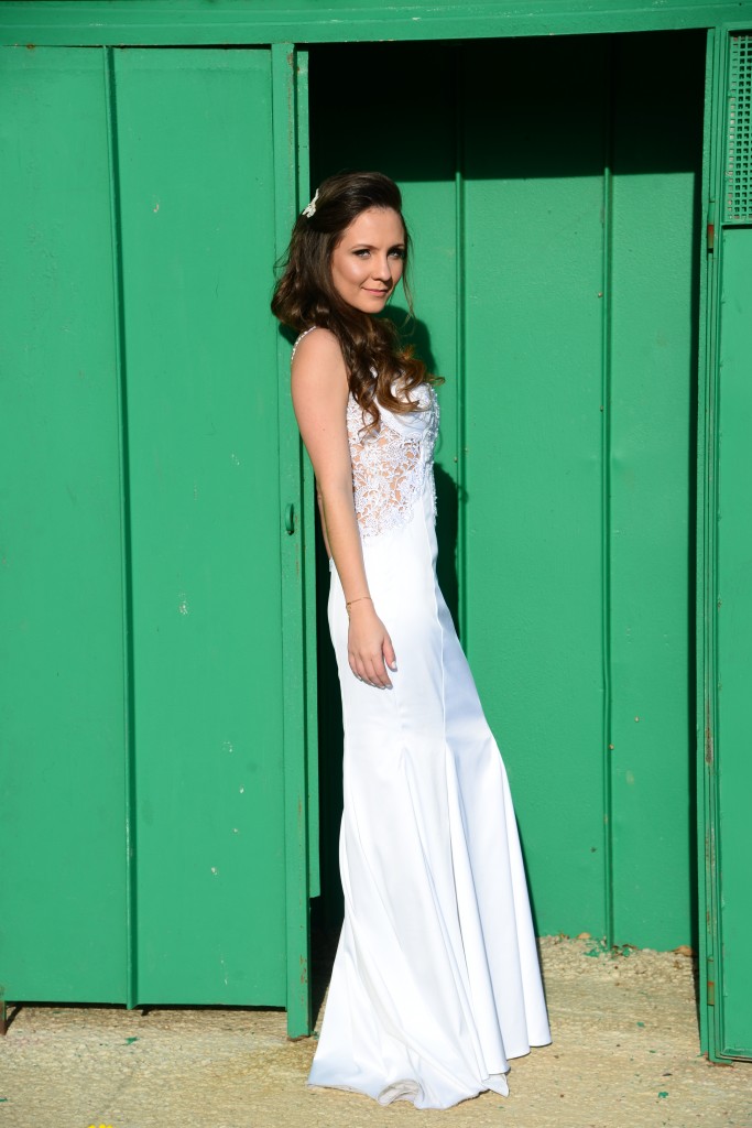 אישה בשמלה לבנה ללא שרוולים עם פרטי תחרה עומדת מול דלת ירוקה ומחייכת. יש לה שיער חום ארוך וגלי והיא מוארת באור השמש, מטיל צל מאחוריה.